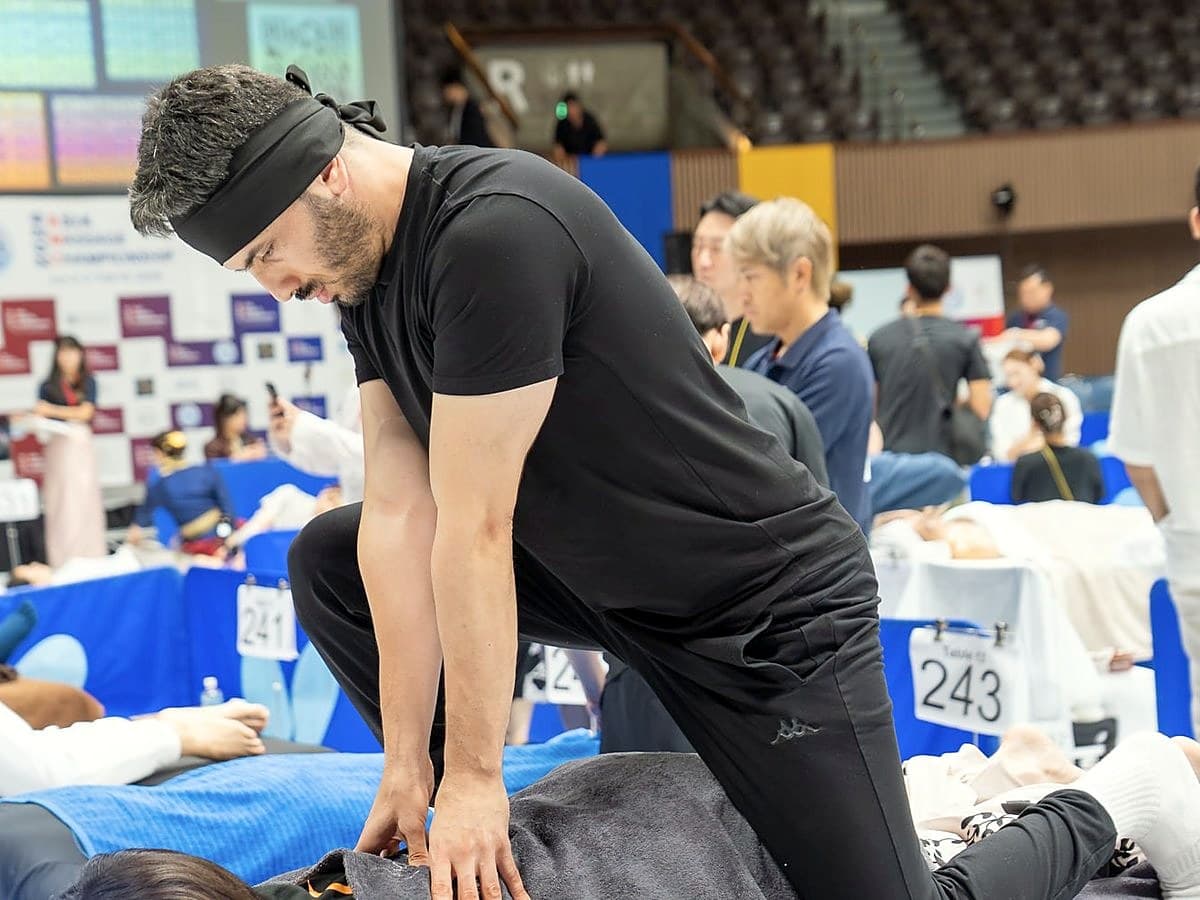 في فئة Body Care ضمن AMC2026، رجل يرتدي قميصًا أسود وعصابة رأس، جاثيًا على ركبة واحدة، يعالج ظهر المتلقي بكلتا يديه